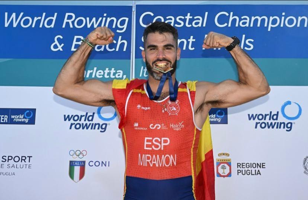 Ein Mann steht auf einem Podium mit erhobenen Armen, trägt eine Medaille und eine Uhr, vor einer Tafel mit Logos und Text.