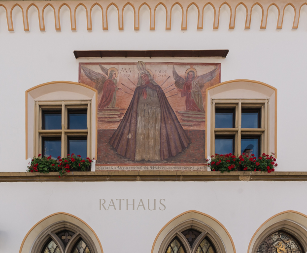 Building with windows and a mural on the side, surrounded by flowering plants, with "Rathaus" text at the bottom.