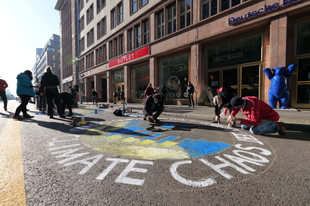 Eine Gruppe von Menschen sitzt vor einem Gebäude mit Fenstern und Namensschildern auf dem Boden, umgeben von Flaschen und anderen Gegenständen, mit Bäumen und einem klaren blauen Himmel im Hintergrund, während sie an einer Klimaprotestaktion in Berlin teilnehmen.