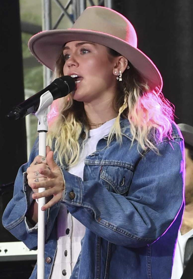 Miley Cyrus performing at the CMA Music Festival in Nashville, Tennessee, singing into a microphone with a stand while wearing a hat, with a person in a cap to her right.
