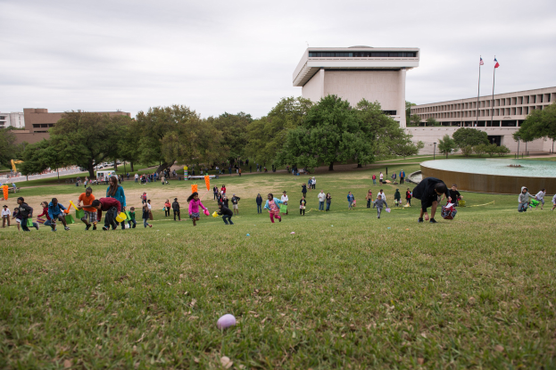 Eine Gruppe von Menschen, die mit Ostereiern auf einer grünen Wiese mit Bäumen, Gebäuden, Fahnen, einem Brunnen und einem bewölkten Himmel spielt.