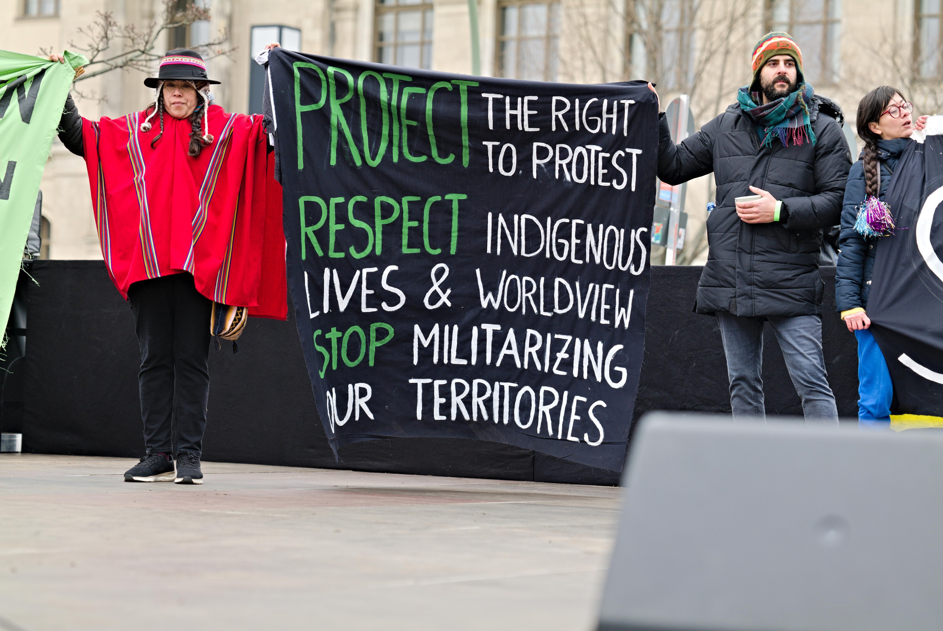 A group of people holding a banner that reads "Protect the Right to Protest Respect Indigenous Lives and Worldview Stop Militarizing Our Territories" in front of a building with trees and a cloudy sky.