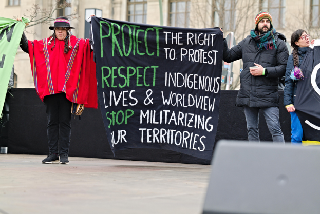 A group of people holding a banner that reads "Protect the Right to Protest Respect Indigenous Lives and Worldview Stop Militarizing Our Territories" in front of a building with trees and a cloudy sky.