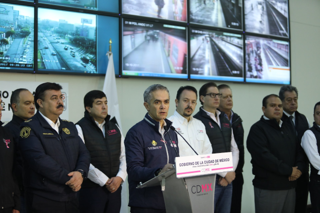 Eine Gruppe von Männern steht vor einem Rednerpult mit einem Mikrofon und einer Tafel, auf der "Mexikanische Zoll- und Grenzschutzbehörde" steht, mit Fernsehgeräten und einer Flagge an der Wand dahinter.