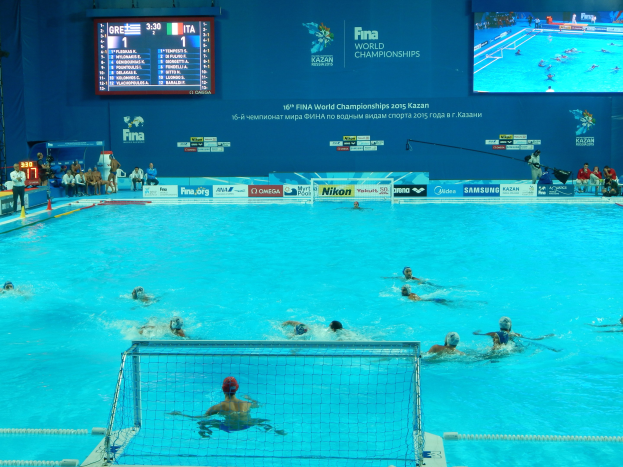 Gruppe von Athleten, die Wasserball in einem Pool während der FINA-Weltmeisterschaften 2016 in Kazan spielen, mit Zuschauern auf der linken Seite und einem Werbeschild im Hintergrund.