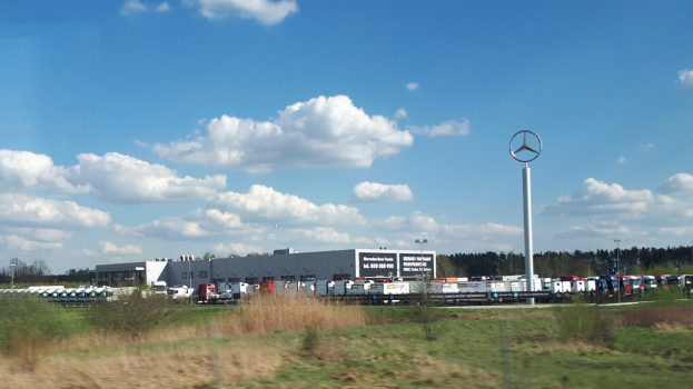 Ein Mercedes-Benz-Werk in Deutschland mit Fahrzeugen auf der Straße, umgeben von Grünflächen, Gebäuden und einem Logo-Mast unter einem bewölkten Himmel.