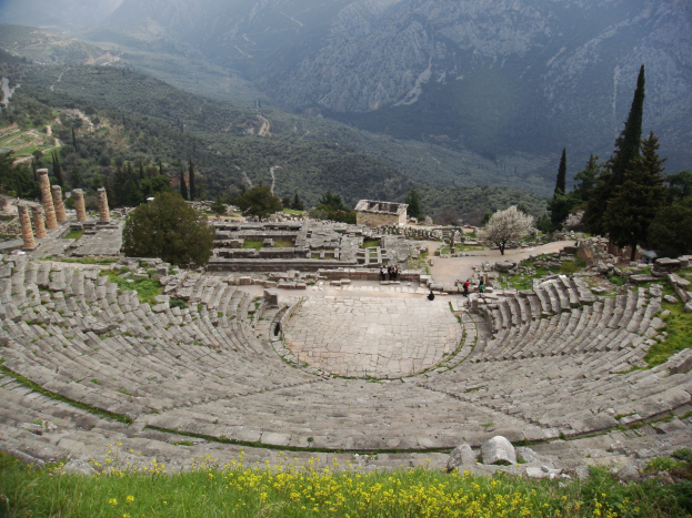Ancient stone theatre in Delphi, Greece, with visitors on stage, surrounded by greenery, ruins, and hills under a blue sky.