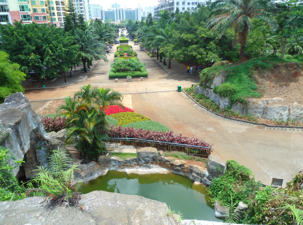 Luftaufnahme eines Parks mit einem Teich, umgeben von Felsen, Pflanzen, Bäumen und Blumen, Menschen, die spazieren gehen, Gebäude im Hintergrund und einem klaren blauen Himmel.