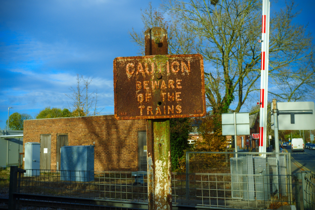 Vorsichtsschild an der Seite einer Bahnschiene, umgeben von einem Metallzaun, mit Bäumen, Strommästen, einem Gebäude, Containern, Fahrzeugen auf einer Straße und einem bewölktem Himmel.
