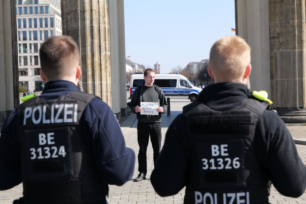 Three police officers stand in front of a building, with a man in the center holding a paper, flanked by pillars and surrounded by vehicles, people, trees, and buildings under a clear sky.