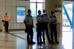 Eine Gruppe von maskierten Polizisten steht vor einem Gebäude mit Glastüren, Schildern mit Text, einer Säule und einem Geländer, wobei durch die Glastüren im Hintergrund Bäume und Fahrzeuge zu sehen sind, was darauf hinweist, dass sie in Hong Kong verhaftet wurden.