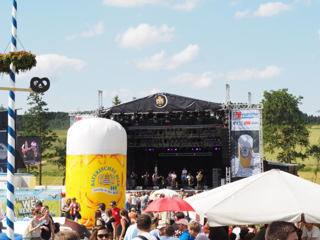Large crowd at Oktoberfest in Munich, Germany, with people in caps and goggles, some holding umbrellas, in front of a stage under a tent with musical performers and a decorated pole.