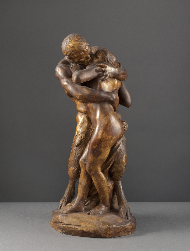 Bronzestatuette namens 'Der Kuss' von Jean-Baptiste-Camille Corot, die einen Mann in einem weißen Hemd und blauen Jeans zeigt, der eine Frau in einem orangefarbenen Kleid gegen einen grauen Hintergrund umarmt.