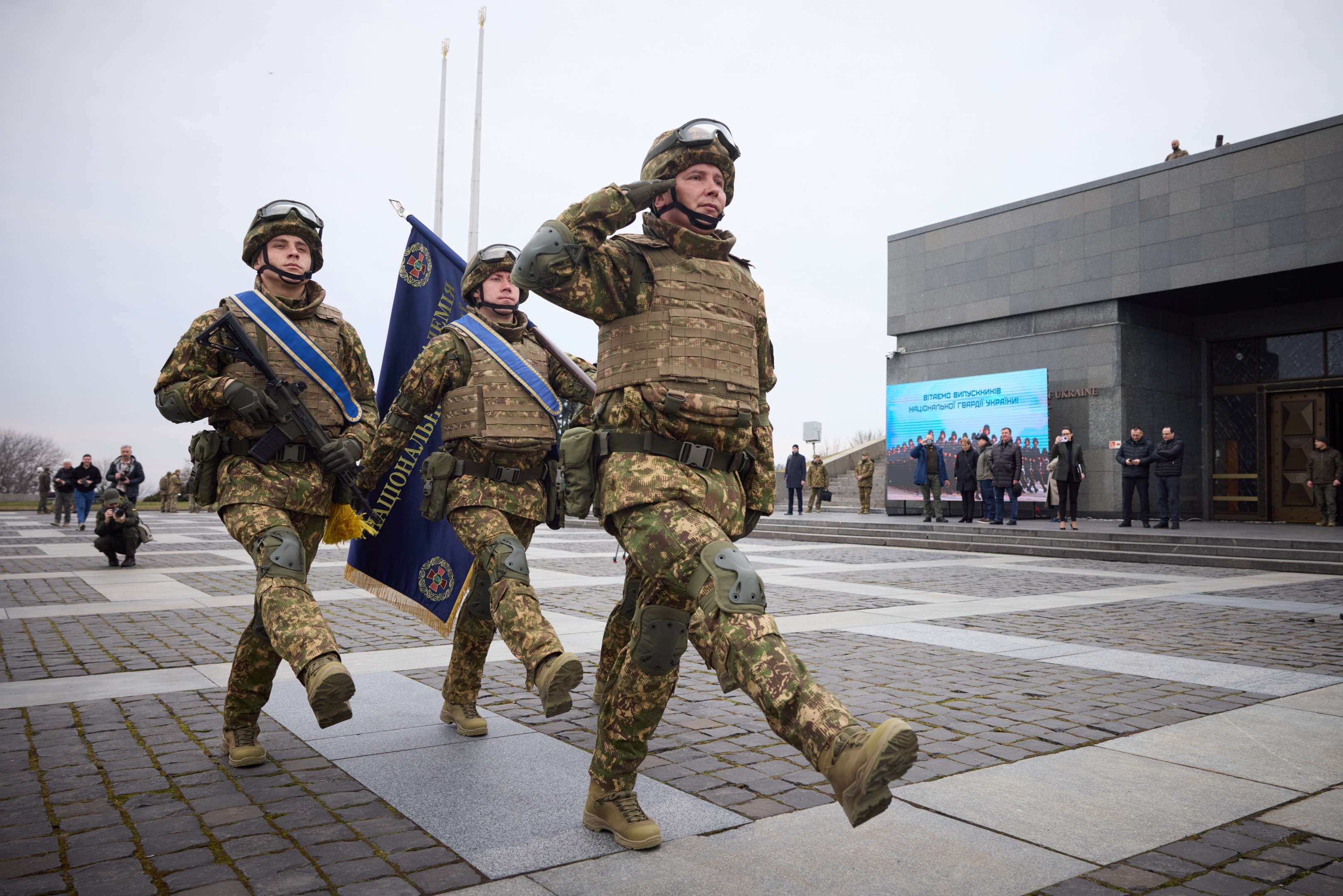 A group of Ukrainian soldiers in uniform march down a street, carrying guns and flags, with a building, people, trees, and a clear sky in the background.