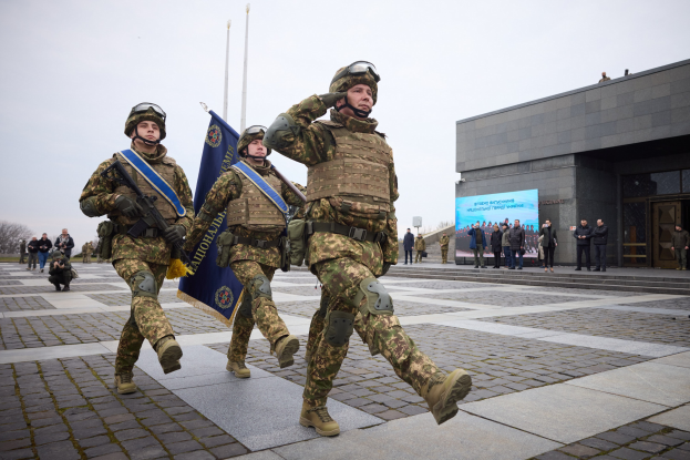 A group of Ukrainian soldiers in uniform march down a street, carrying guns and flags, with a building, people, trees, and a clear sky in the background.