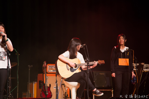 Drei Mädchen auf der Bühne, zwei singen in Mikrofone und eines hält ein Musikinstrument, mit einer Gitarre, einem Lautsprecher und einer Schalttafel.