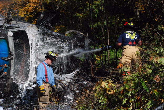 Zwei Feuerwehrleute in Helmen und Handschuhen, einer sprüht Wasser aus einer Rohrleitung auf ein umgestürztes Auto im Wald, mit Bäumen und Maschendrahtzaun im Hintergrund.