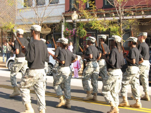 Mehrere Personen in schwarzen Oberteilen und Kappen sind auf einer Straße unterwegs, einige halten Waffen, mit einem Fahrzeug in der Nähe; einige Menschen sind auf dem Gehweg, eine Straße ist in der Mitte, und es gibt Trees und Gebäude im Hintergrund.