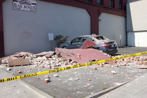 Eine eingestürzte Wand mit verstreuten roten Ziegeln, ein geparktes Auto, eine Topfpflanze, nahe gelegene Gebäude und eine Werbetafel mit der Aufschrift "Kautionsfreiheit".