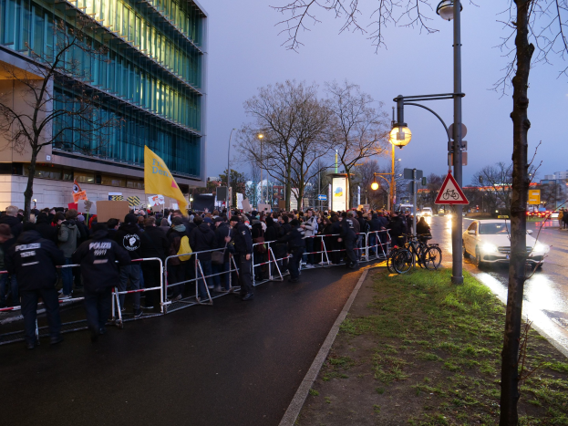 Eine große Gruppe von Menschen mit Schildern steht vor einem Gebäude, mit Barrikaden, Fahrrädern, Laternen, Schildern, Bäumen und Gras im Vordergrund und dem Himmel im Hintergrund, was auf eine Protestaktion in Berlin hinweist.