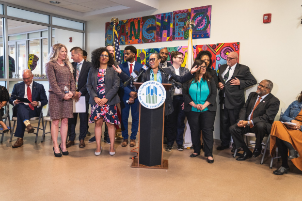 Eine Gruppe von Menschen, die vor einem Podium mit einem Mikrofon und einer Tafel mit Text darauf stehen und sitzen, mit Flaggen, einem Feuerlöscher und Deckenlampen an der Rückwand.