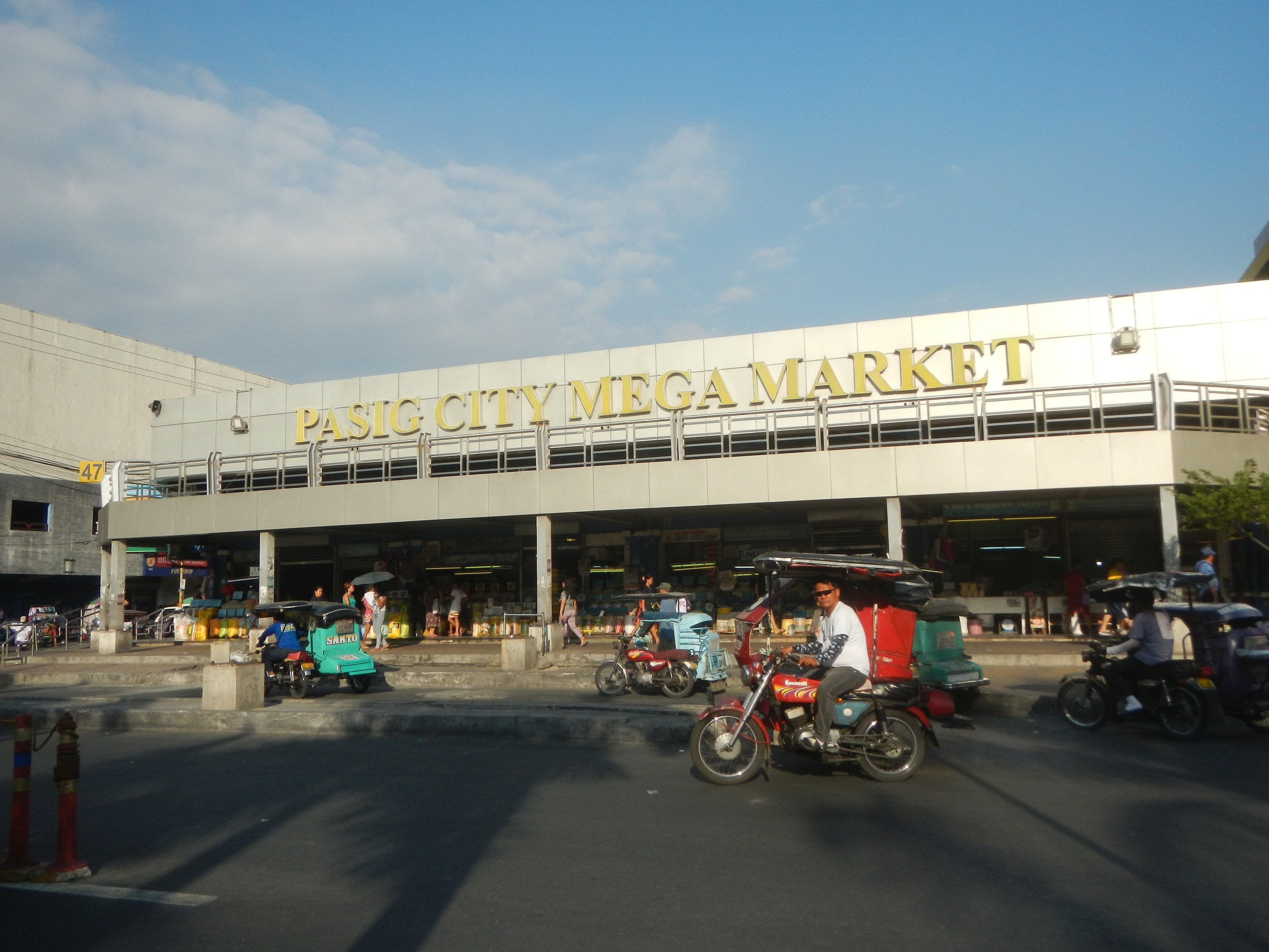 Eine belebte Straßenszene am Pasig City Mega Market mit Fahrzeugen und Passanten, ein Gebäude mit Text im Hintergrund und ein bewölkter Himmel.