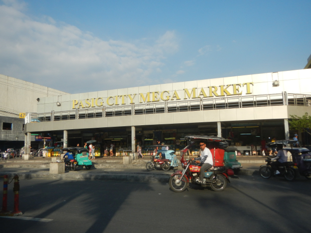 Eine belebte Straßenszene am Pasig City Mega Market mit Fahrzeugen und Passanten, ein Gebäude mit Text im Hintergrund und ein bewölkter Himmel.