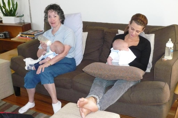 Zwei Frauen sitzen auf einem Sofa und halten Babys, mit Kissen drumherum, vor einer wei\374en Wand, mit einem Schreibtisch, B"chern und einer Zimmerpflanze links daneben.