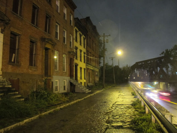 Eine Stadtansicht mit Gebäuden, Treppen mit einem Metallzaun, Strommasten, Laternenmasten mit Kabeln, einer Straße, einem Metallzaun, einer Brücke, darunter fahrende Autos und Laternenmasten auf dem Gehweg.