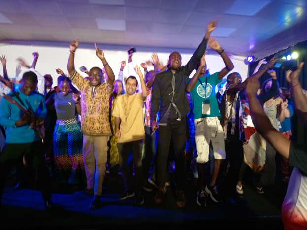 Eine Gruppe von Menschen, die auf einer Bühne tanzen und die Hände heben, beleuchtet von Scheinwerfern auf der rechten Seite und einem Hintergrundbild.