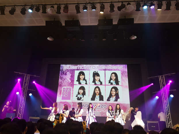 Eine Gruppe von Menschen, die auf einer hell erleuchteten Bühne mit einer Menge darunter performen, mit einem Sänger mit Mikrofon und einem Musiker, der ein Instrument spielt, während ein großer Bildschirm Bilder der K-Pop-Gruppe BK48 anzeigt.