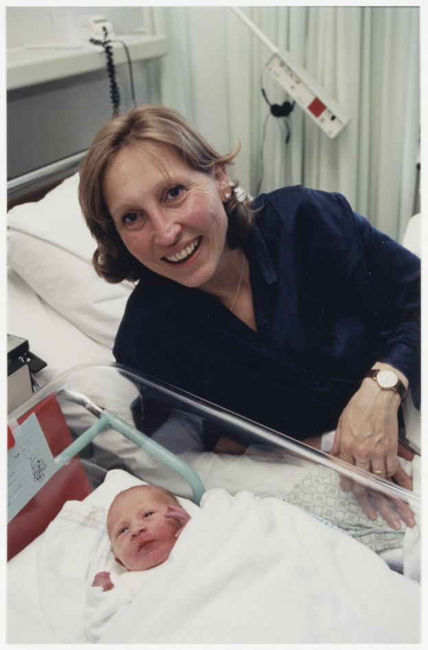 Eine Frau in einem blauen Kleid und Uhr hält ein Baby im Arm, während sie in einem Krankenhausbett liegt, umgeben von medizinischer Ausrüstung und einem weißen Vorhang.