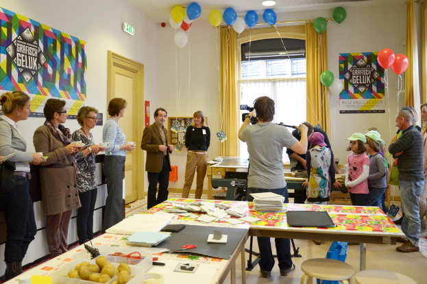 Ein voller Raum mit feiernden Menschen, Tischen mit verschiedenen Gegenständen wie Büchern und Kartoffeln, einem Mann, der mit einer Kamera auf einem Stativ filmt, Menschen mit Tassen in der Hand und Kindern in der Ecke mit Mützen, sowie einem Fenster und Vorhängen im Hintergrund.