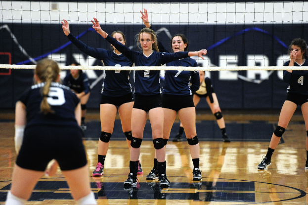 Drei Mädchen bei einem Volleyballspiel auf dem Feld stehend mit erhobenen Händen, ein Netz vor ihnen und ein Zaun im Hintergrund.