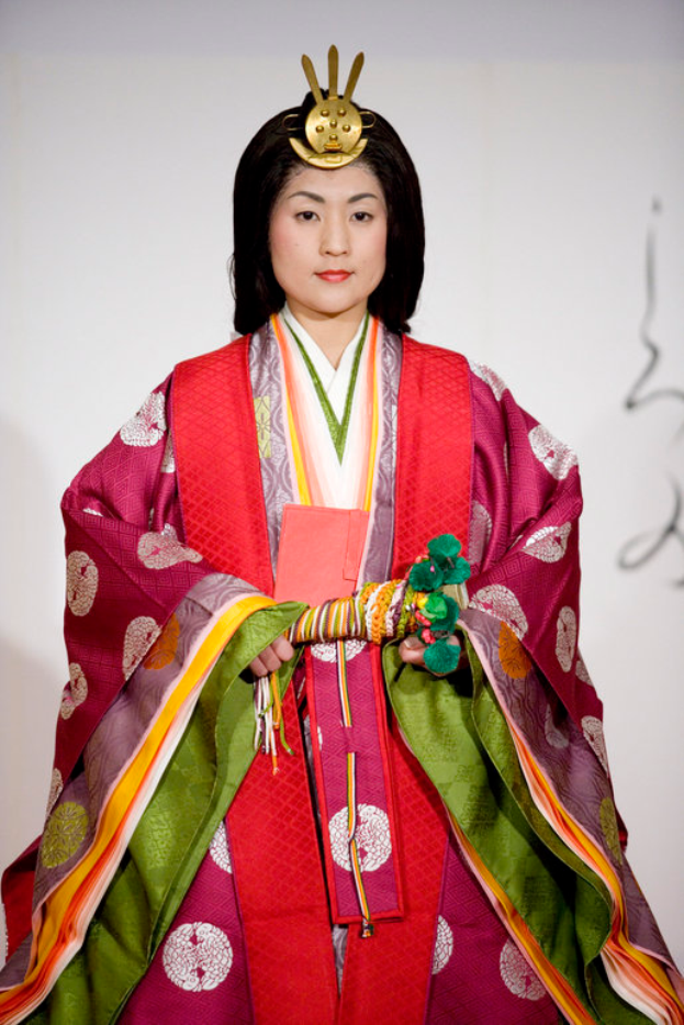 Eine Frau in einem traditionellen japanischen Kimono, das ein leuchtend rotes und grünes Kleid mit aufwändigen Mustern trägt, steht selbstbewusst auf einem Laufsteg, ihr Haar ordentlich zu einem Knoten gebunden.