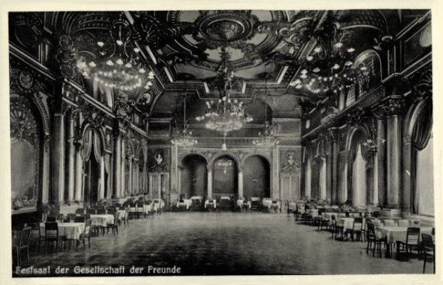 Black and white photo of a large room with tables, chairs, pillars, arches, and ceiling chandeliers, labeled "Festival der Gesellschaft der Freunde, Berlin" at the bottom.