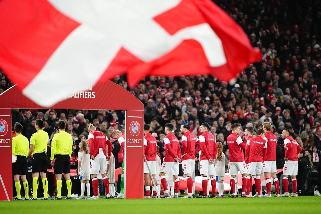 Eine Gruppe von Menschen auf einem Fußballfeld mit einer roten und weißen Flagge im Vordergrund, einem Bogen mit der Aufschrift "Bayern München vs. Bayern München Wetten & Vorschau" im Hintergrund und einer großen Menge im Stadion.