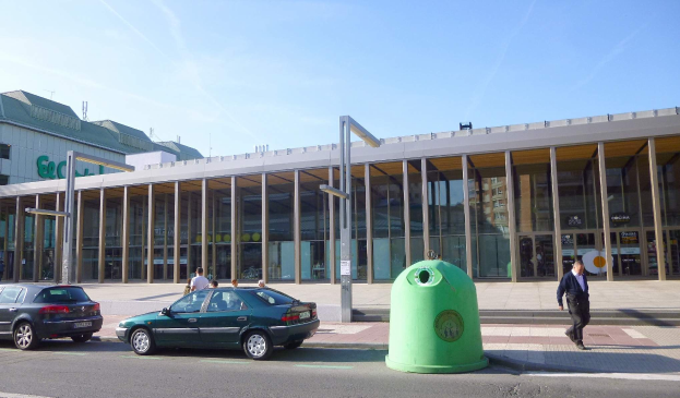 Grüner Müllcontainer auf einer Stadtstraße neben einem Glaswandgebäude mit Säulen, mit Fahrzeugen und Fußgängern im Vordergrund und einer bewölkten Himmel im Hintergrund, am Berliner Hauptbahnhof.