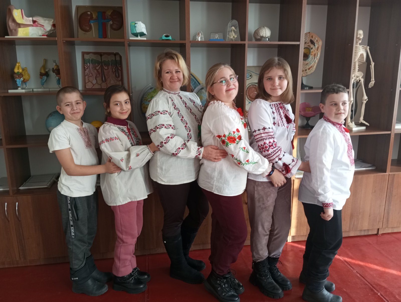 A group of children in traditional Ukrainian clothing standing in front of a bookshelf with toys, a skeleton, and other objects.