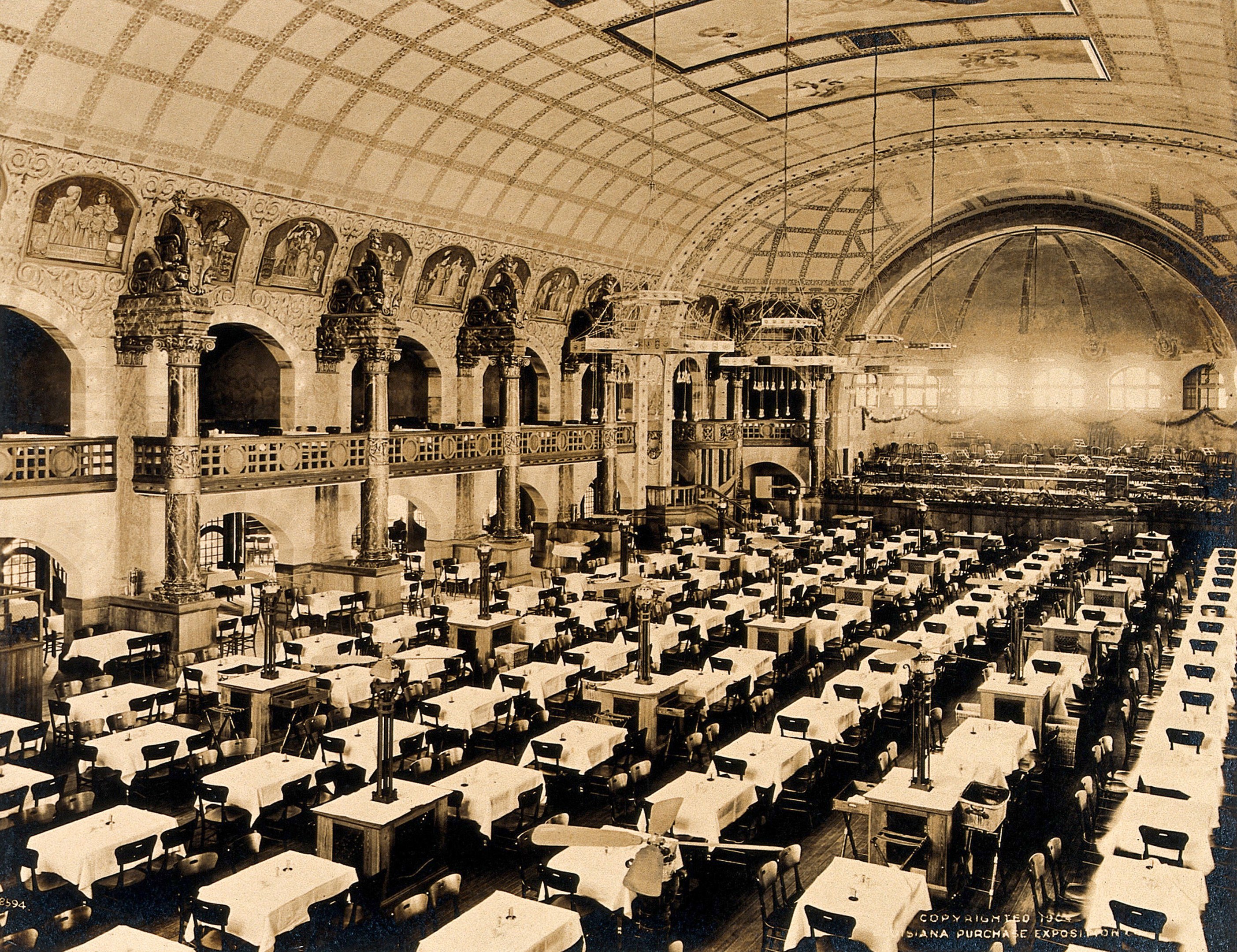 A vintage photograph of a large, ornate room with numerous tables and chairs, decorative pillars, wall-mounted sculptures, and ceiling lights.