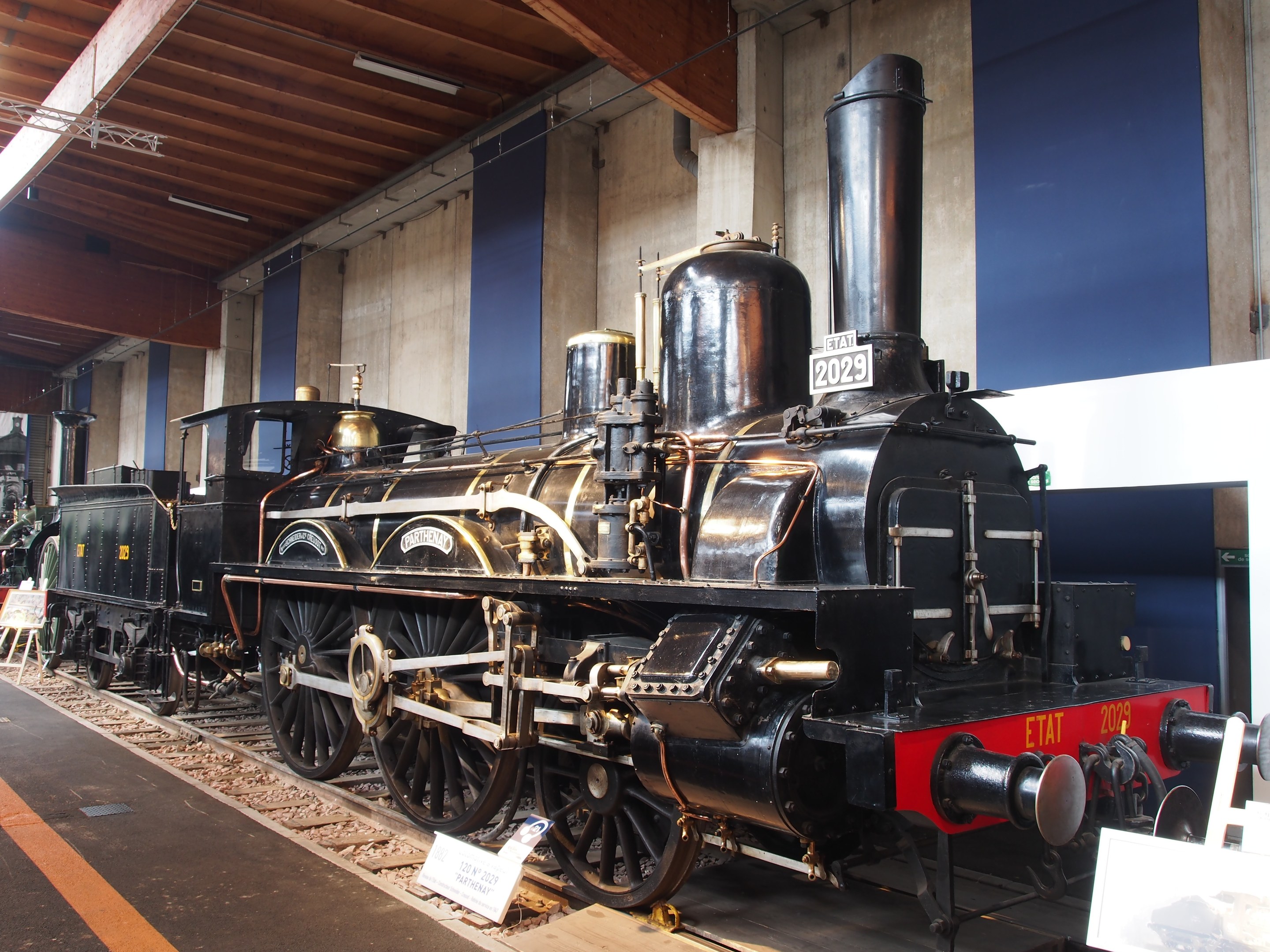 Alte Dampflokomotive in einem Museum ausgestellt, umgeben von Eisenbahnschienen und informativen Schildern, mit einer Wand und einer beleuchteten Decke im Hintergrund.