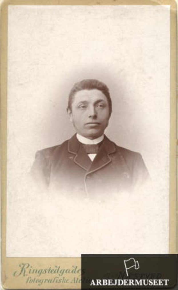 Ein altes Schwarz-Weiß-Foto eines Mannes in einem dunklen Anzug, weißem Hemd und schwarzer Krawatte, mit ordentlich frisiertem Haar, der direkt in die Kamera schaut und ein ernstes Gesicht mit einem leichten Grinsen hat.