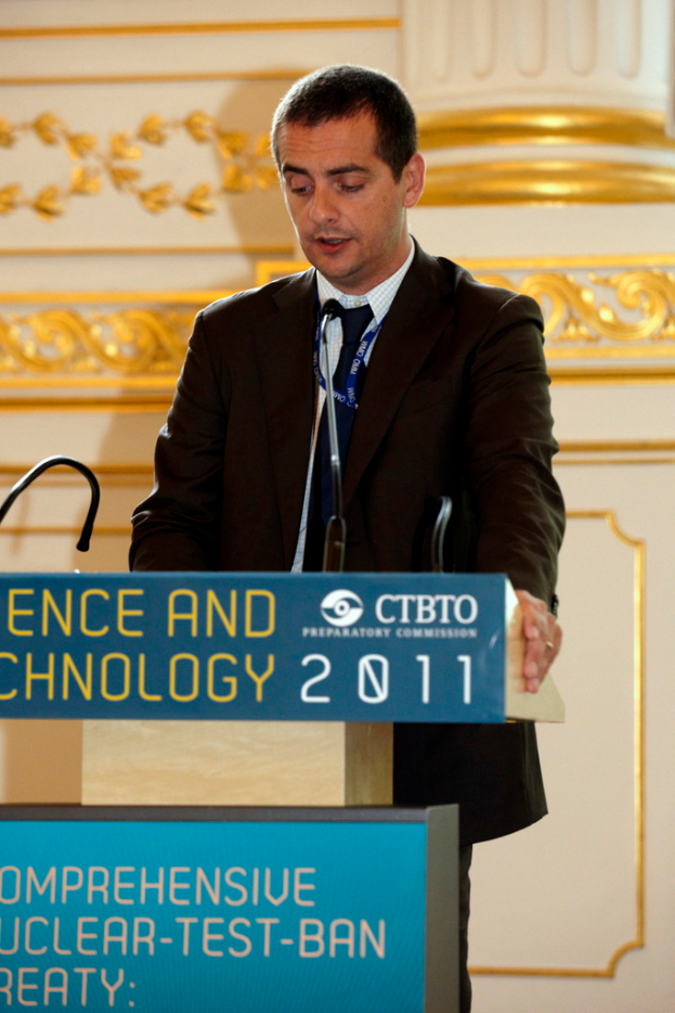 Ein Mann in einem Anzug steht neben einem Rednerpult mit einer Maus und Plakaten darauf, mit einer Säule und einer Wand im Hintergrund.