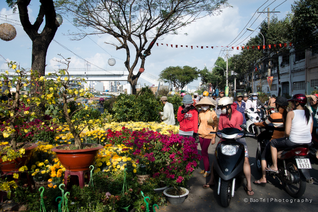 Eine Gruppe von Menschen in Helmen und MÃ¼tzen, einige zu Fuss und einige mit dem Fahrrad auf einer Strasse, mit Pflanzen, einem Blumentopf auf einem Hocker, BÃ¤umen, einem Pfahl, DrÃ¤hten, einer Lampe, einer BrÃ¼cke und GebÃ¤uden in der NÃ¤he.