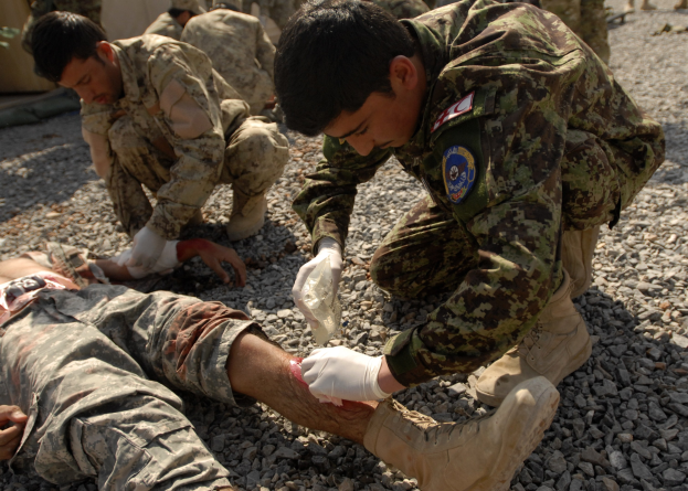 A group of men in military uniforms kneeling on the ground, with one lying on the stones, one wearing a bandage on his leg, and others standing in the background near a tent.