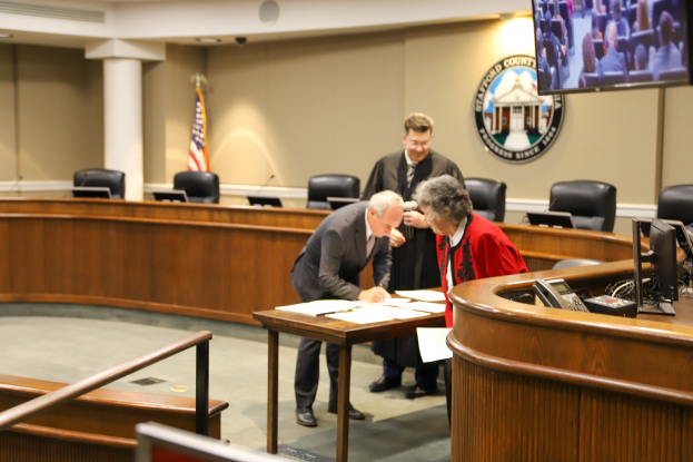 Eine Gruppe von drei Personen, die um einen Tisch in einem Gerichtssaal stehen und Papiere unterschreiben, mit verschiedenen Gegenständen auf dem Tisch und Elementen wie Stühlen, einer Flagge, einem Logo, einem Fernseher und Deckenleuchten im Hintergrund.