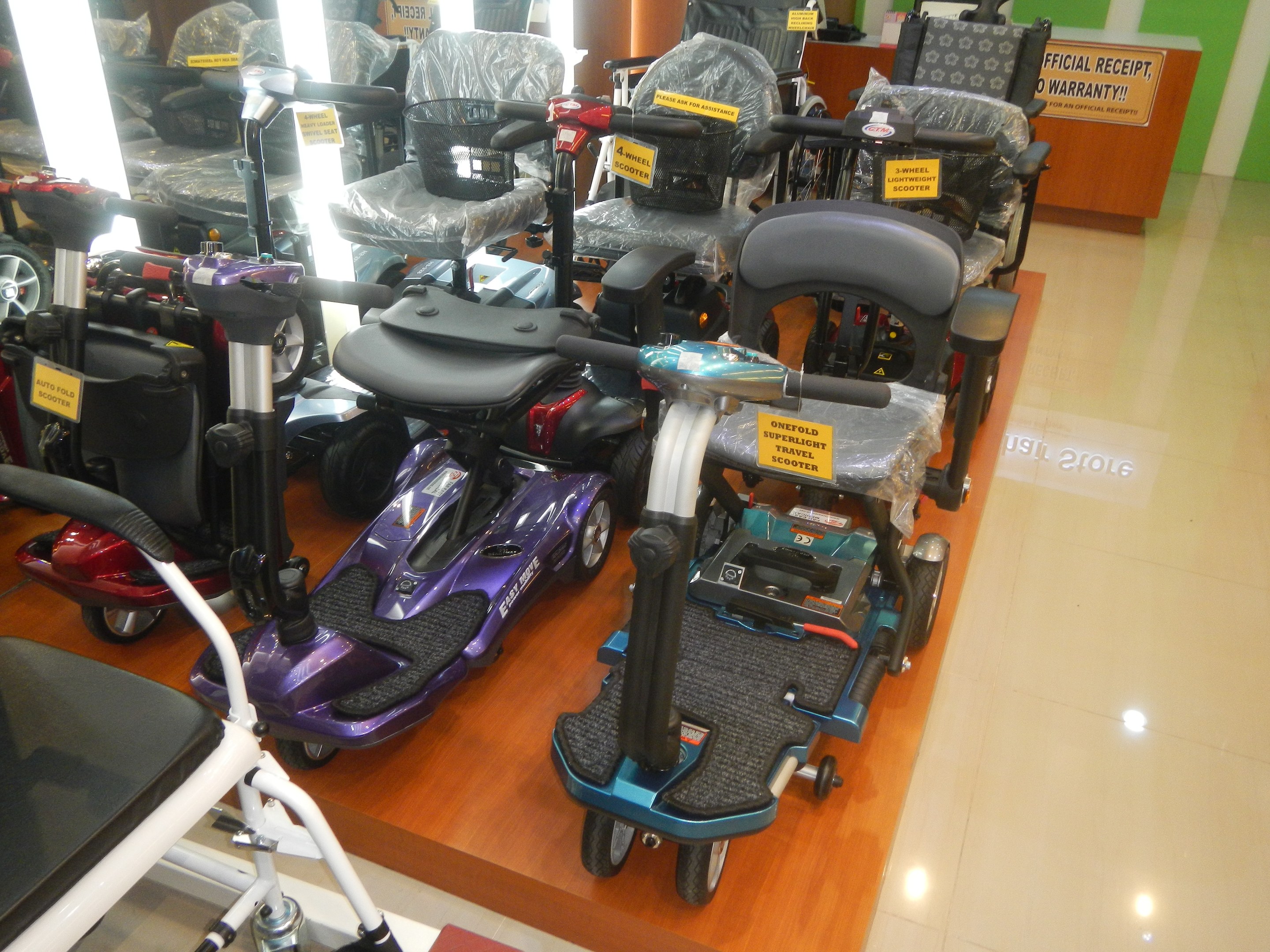 Eine Reihe von Elektro-Scootern auf einer Verkaufsfläche mit Preisschildern, einem Holztisch mit einer Tafel im Hintergrund und sichtbarem Boden.
