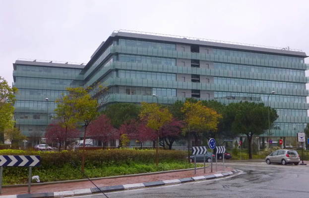 Großes modernes Gebäude mit zahlreichen Fenstern, der Hauptsitz der Europäischen Zentralbank, umgeben von Grünflächen, Fahrzeugen, Straßeninfrastruktur und einem bewölkten Himmel.