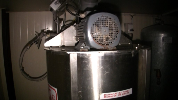 Ein Stahlfass mit Motorkabeln und anderen Gegenständen darauf, ein weiteres Fass in der Nähe einer Tür und Aufkleber unten.