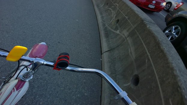 Unten ist ein Fahrradlenker zu sehen, mit Fahrzeugen auf der rechten Straße und einer Mittellinie in der Mitte.